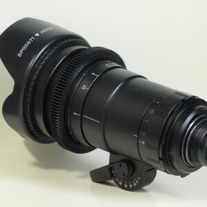 May include: A black camera lens with a lens hood. The lens has a series of markings and a gear ring. The lens hood has the text "SPT-W077" and an arrow pointing down.