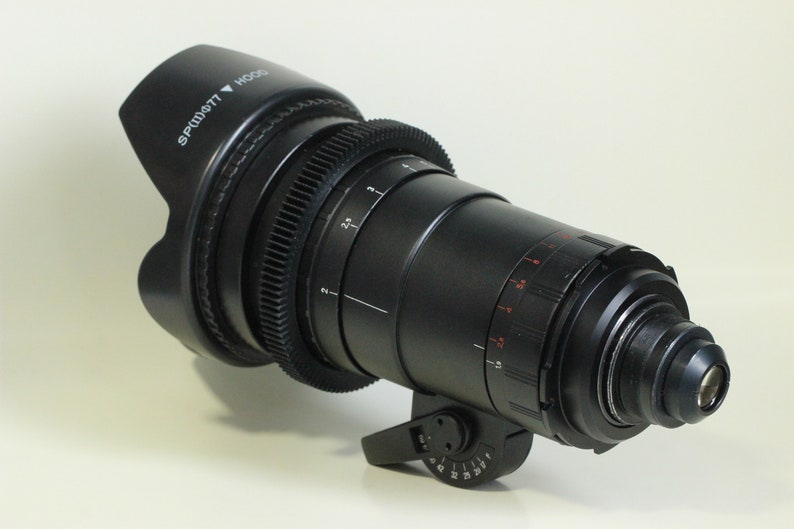 May include: A black camera lens with a lens hood. The lens has a series of markings and a gear ring. The lens hood has the text "SPT-W077" and an arrow pointing down.
