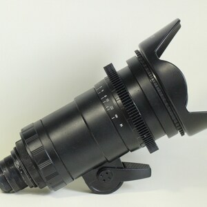 May include: A black camera lens with a lens hood. The lens has a focus ring and a gear ring. The lens hood is made of black plastic and has a rectangular shape.