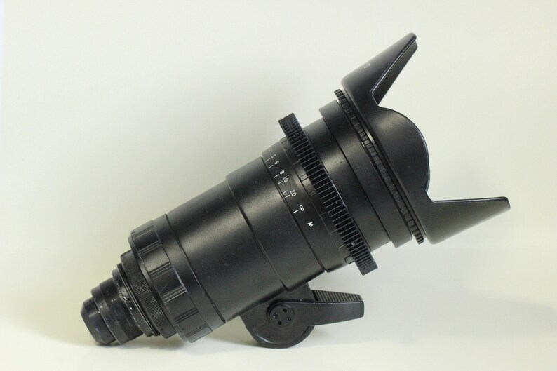 May include: A black camera lens with a lens hood. The lens has a focus ring and a gear ring. The lens hood is made of black plastic and has a rectangular shape.