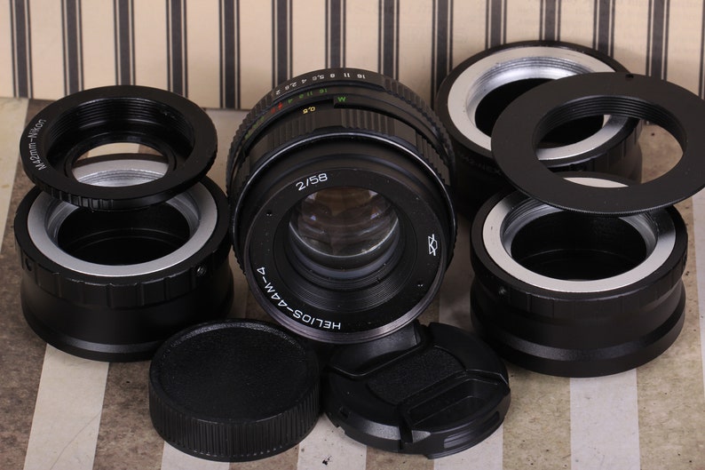 Pu&ograve; includere: Un obiettivo per fotocamera nero con un anello argentato e il testo "HELIOS-44M-4" e "2/58" stampato sul lato. L'obiettivo &egrave; circondato da diversi adattatori per obiettivi per fotocamera neri e argentati.