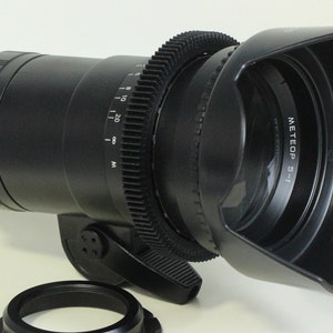 May include: A black camera lens with a lens hood and a gear ring. The lens has a depth of field scale marked in meters and feet. The lens hood has the text "METEOP 5-1" printed on it.