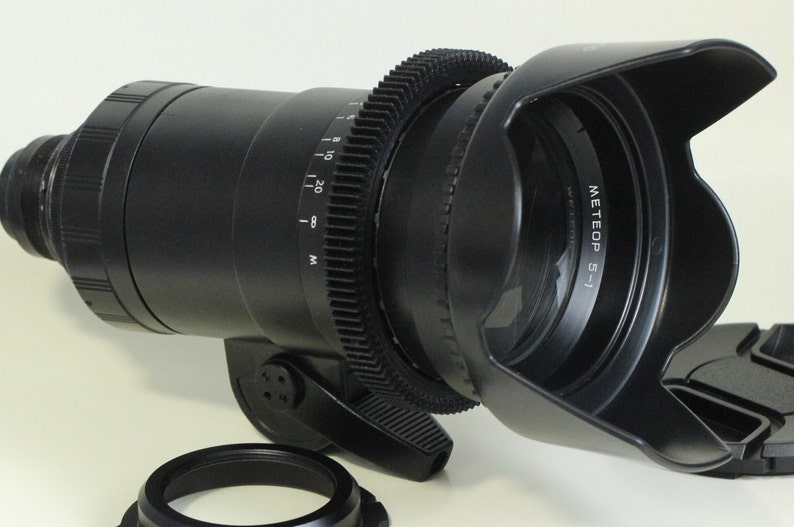 May include: A black camera lens with a lens hood and a gear ring. The lens has a depth of field scale marked in meters and feet. The lens hood has the text "METEOP 5-1" printed on it.