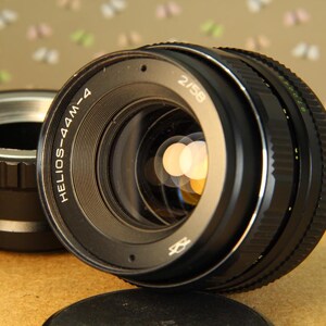 Pode incluir: Uma lente de c&acirc;mera preta com o texto "HELIOS-44M-4 2/58" impresso na lateral. A lente tem um anel prateado ao redor da frente e uma tampa de metal preta.