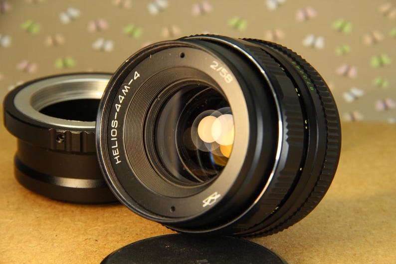 Pode incluir: Uma lente de c&acirc;mera preta com o texto "HELIOS-44M-4 2/58" impresso na lateral. A lente tem um anel prateado ao redor da frente e uma tampa de metal preta.