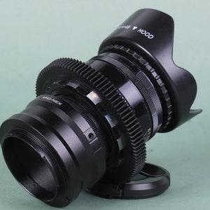 Puede incluir: Un objetivo de c&aacute;mara negro con parasol y anillos de engranajes. El objetivo tiene el texto "M42-NEX" y "HOOD" impreso. El objetivo est&aacute; ligeramente inclinado hacia la derecha sobre un fondo verde claro.