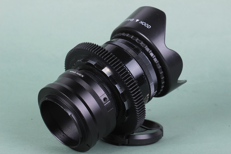 Puede incluir: Un objetivo de c&aacute;mara negro con parasol y anillos de engranajes. El objetivo tiene el texto "M42-NEX" y "HOOD" impreso. El objetivo est&aacute; ligeramente inclinado hacia la derecha sobre un fondo verde claro.
