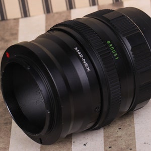 Pu&ograve; includere: Un obiettivo per fotocamera nero con un anello argentato e il testo "M42-NEX" e "830051" stampato su di esso.