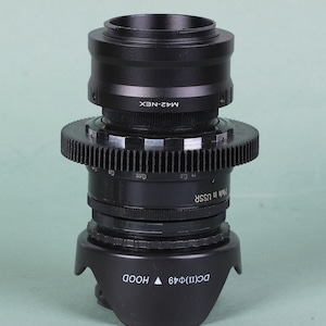 Puede incluir: Adaptador de lente de c&aacute;mara negro con un anillo en forma de engranaje y un parasol. El adaptador est&aacute; etiquetado como "M42-NEX" y "Made in USSR". El parasol est&aacute; etiquetado como "DC(II)&Phi;49 HOOD".