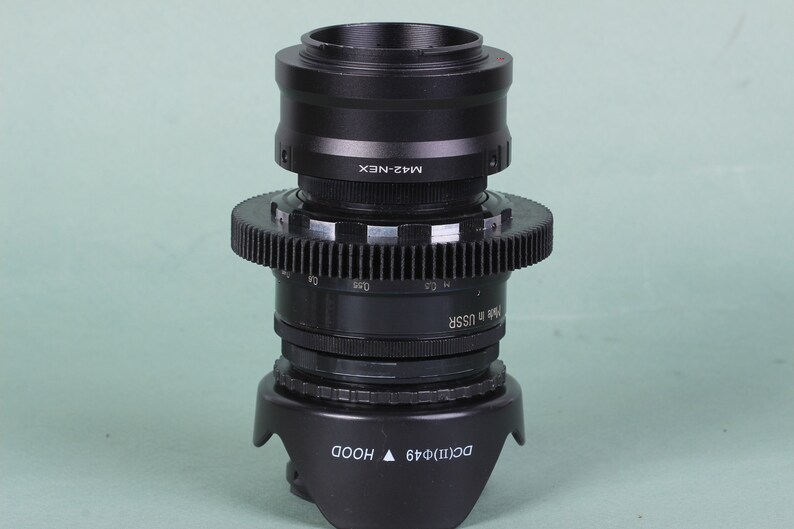 Puede incluir: Adaptador de lente de c&aacute;mara negro con un anillo en forma de engranaje y un parasol. El adaptador est&aacute; etiquetado como "M42-NEX" y "Made in USSR". El parasol est&aacute; etiquetado como "DC(II)&Phi;49 HOOD".
