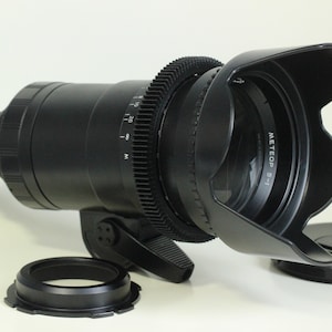 May include: A black camera lens with a lens hood and a gear ring. The lens hood has the text "METEOP 5-1" printed on it.