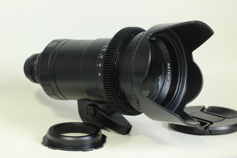 May include: A black camera lens with a lens hood and a gear ring. The lens hood has the text "METEOP 5-1" printed on it.