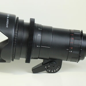 May include: A black camera lens with a lens hood. The lens has a gear ring and a focus ring. The lens hood has the text "SPLD 077" printed on it.