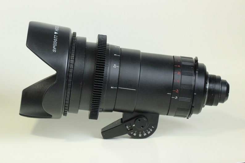 May include: A black camera lens with a lens hood. The lens has a gear ring and a focus ring. The lens hood has the text "SPLD 077" printed on it.