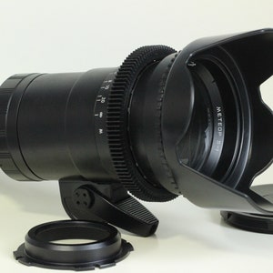 May include: A black camera lens with a lens hood attached. The lens has a gear ring and a focus ring. The lens hood is made of black plastic and has a black rubber ring around the edge. The lens is sitting on a white surface.