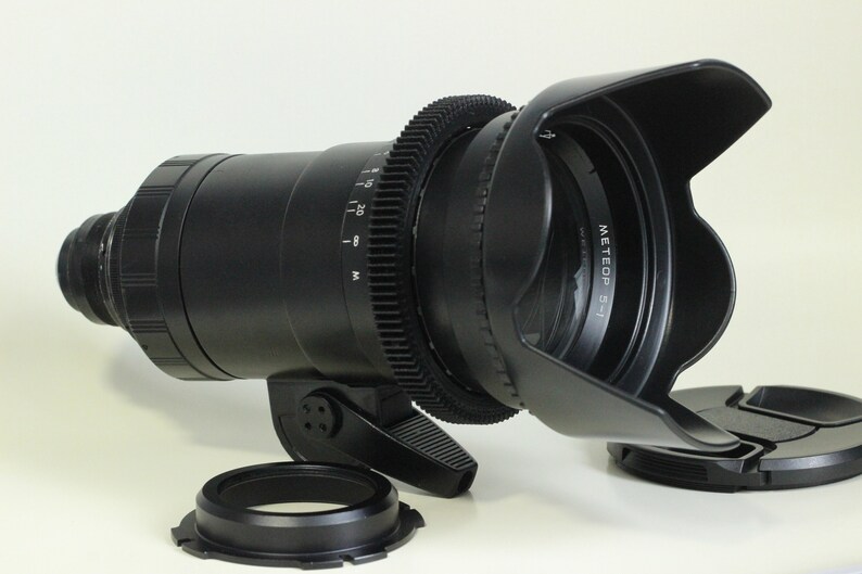May include: A black camera lens with a lens hood attached. The lens has a gear ring and a focus ring. The lens hood is made of black plastic and has a black rubber ring around the edge. The lens is sitting on a white surface.