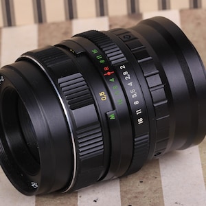 Pu&ograve; includere: Un obiettivo per fotocamera nero con un anello argentato e un tappo dell'obiettivo nero. L'obiettivo ha una scala di profondit&agrave; di campo con segni per le aperture da f/2,8 a f/16. L'obiettivo ha anche una scala di distanza con segni in metri e piedi.