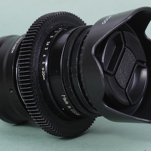 Puede incluir: Un objetivo de c&aacute;mara negro con parasol y tapa de objetivo. El objetivo tiene un anillo en forma de engranaje y marcas para el enfoque y la apertura. El texto "Made in USA" es visible.