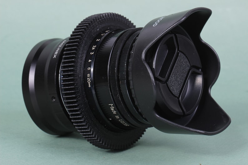 Puede incluir: Un objetivo de c&aacute;mara negro con parasol y tapa de objetivo. El objetivo tiene un anillo en forma de engranaje y marcas para el enfoque y la apertura. El texto "Made in USA" es visible.