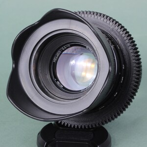 Puede incluir: Un objetivo de c&aacute;mara negro con parasol incorporado. El objetivo tiene un anillo de enfoque estriado y un elemento de vidrio transparente que refleja la luz. El objetivo est&aacute; montado sobre una base negra y sobre un fondo verde claro.