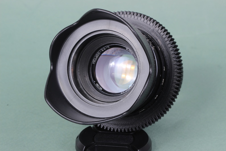 Puede incluir: Un objetivo de c&aacute;mara negro con parasol incorporado. El objetivo tiene un anillo de enfoque estriado y un elemento de vidrio transparente que refleja la luz. El objetivo est&aacute; montado sobre una base negra y sobre un fondo verde claro.