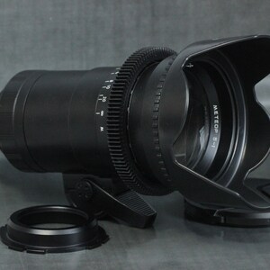 May include: A black camera lens with a lens hood and a gear ring. The lens has a white label that reads "METEOP 5-1".