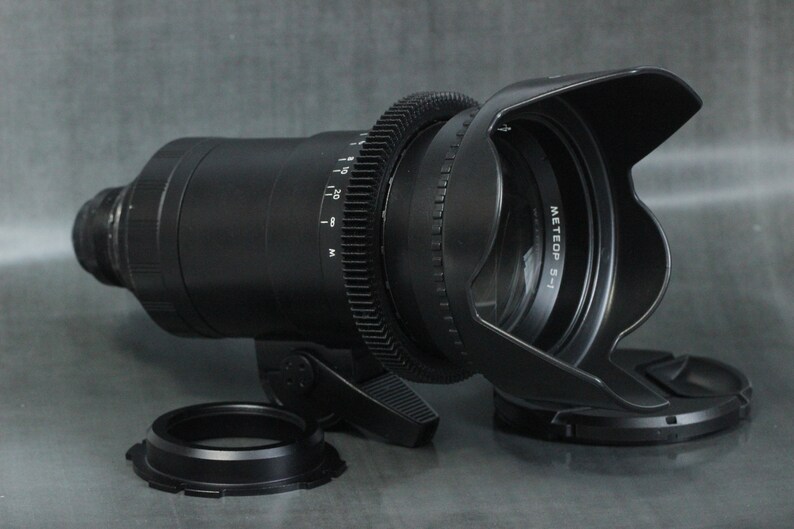 May include: A black camera lens with a lens hood and a gear ring. The lens has a white label that reads "METEOP 5-1".