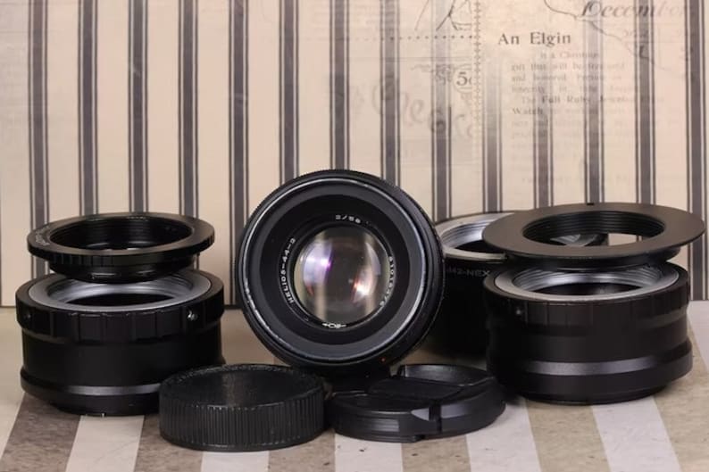 Pu&ograve; includere: Un set di cinque adattatori per obiettivi per fotocamera neri. Gli adattatori sono in metallo e hanno una variet&agrave; di dimensioni. Gli adattatori sono disposti su una superficie a righe.