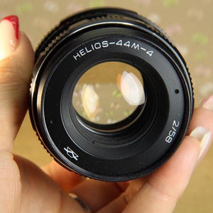Pode incluir: Uma lente de c&acirc;mera preta com o texto "HELIOS-44M-4 2/58" impresso na lateral. A lente tem um anel prateado ao redor da borda e uma lente de vidro transparente.