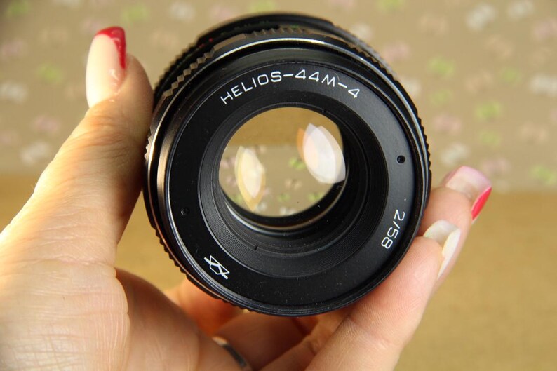 Pode incluir: Uma lente de c&acirc;mera preta com o texto "HELIOS-44M-4 2/58" impresso na lateral. A lente tem um anel prateado ao redor da borda e uma lente de vidro transparente.