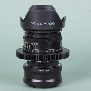 Puede incluir: Un objetivo de c&aacute;mara negro con un anillo en forma de engranaje y un parasol. El objetivo tiene marcas y n&uacute;meros, incluyendo "DC(II)&Phi;49 HOOD" y "M42-NEX". Dise&ntilde;ado para fotograf&iacute;a y videograf&iacute;a.