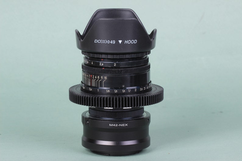 Puede incluir: Un objetivo de c&aacute;mara negro con un anillo en forma de engranaje y un parasol. El objetivo tiene marcas y n&uacute;meros, incluyendo "DC(II)&Phi;49 HOOD" y "M42-NEX". Dise&ntilde;ado para fotograf&iacute;a y videograf&iacute;a.