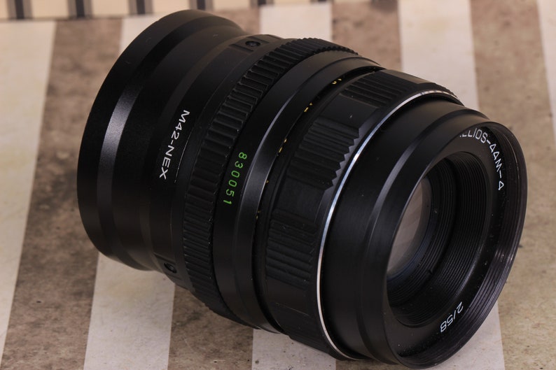 Pu&ograve; includere: Un obiettivo per fotocamera nero con un anello argentato e il testo "Helios-44M-4" e "M42-NEX" stampato su di esso. L'obiettivo ha un numero di serie "830051" stampato su di esso.
