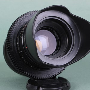 Puede incluir: Un objetivo de c&aacute;mara negro con parasol incorporado. El objetivo tiene un anillo en forma de engranaje en el centro y una serie de n&uacute;meros y marcas. El objetivo est&aacute; ligeramente inclinado, revelando el cristal reflectante del interior. El objetivo est&aacute; sobre una base negra.