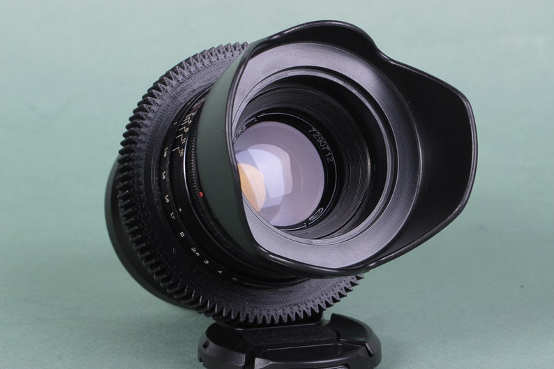 Puede incluir: Un objetivo de c&aacute;mara negro con parasol incorporado. El objetivo tiene un anillo en forma de engranaje en el centro y una serie de n&uacute;meros y marcas. El objetivo est&aacute; ligeramente inclinado, revelando el cristal reflectante del interior. El objetivo est&aacute; sobre una base negra.