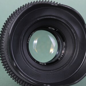 Puede incluir: Primer plano de una lente de c&aacute;mara negra Helios-44-2 con un anillo de enfoque estriado. La lente tiene un elemento de vidrio transparente y el texto "2/58" es visible. La lente est&aacute; sobre un fondo verde claro.