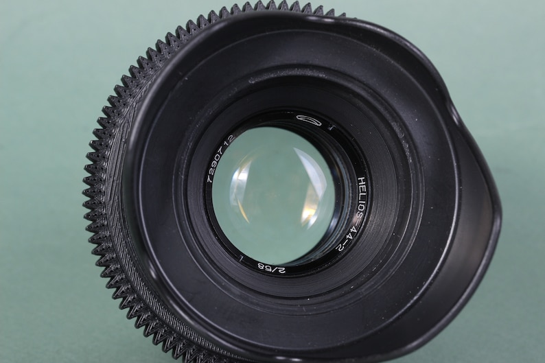 Puede incluir: Primer plano de una lente de c&aacute;mara negra Helios-44-2 con un anillo de enfoque estriado. La lente tiene un elemento de vidrio transparente y el texto "2/58" es visible. La lente est&aacute; sobre un fondo verde claro.