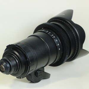 May include: A black camera lens with a lens hood. The lens has a focus ring and a zoom ring. The lens is designed for professional video and film production.