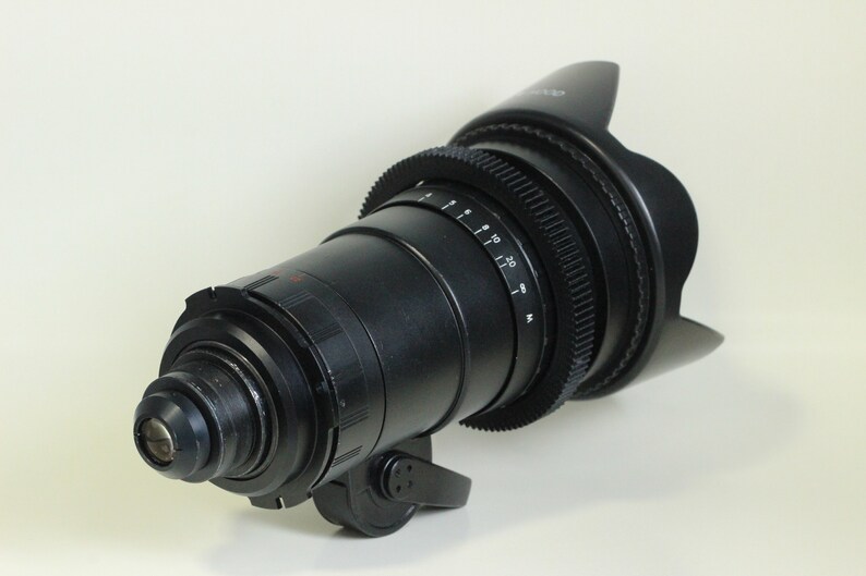 May include: A black camera lens with a lens hood. The lens has a focus ring and a zoom ring. The lens is designed for professional video and film production.