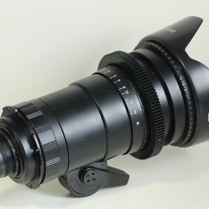 May include: A black camera lens with a lens hood. The lens has a gear ring and a focus ring. The lens hood is made of black plastic and has a rubberized grip.
