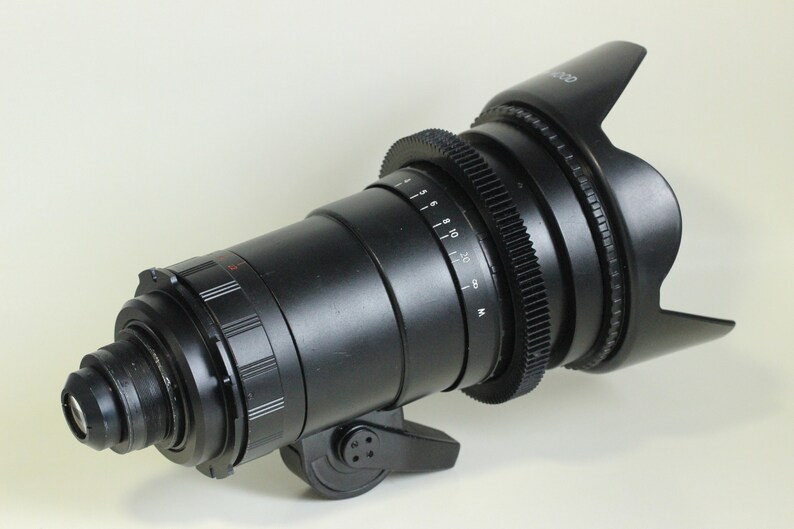 May include: A black camera lens with a lens hood. The lens has a gear ring and a focus ring. The lens hood is made of black plastic and has a rubberized grip.