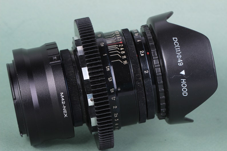 Puede incluir: Un objetivo de c&aacute;mara negro con parasol, con varias marcas de medici&oacute;n y el texto "DC(II)&Phi;49 HOOD". El objetivo tiene un anillo de enfoque y ajustes de apertura, con el texto "M42-NEX" visible.