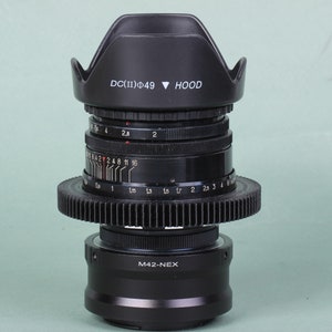 Puede incluir: Un objetivo de c&aacute;mara negro con un parasol con la inscripci&oacute;n "DC(II)&Phi;49 HOOD". El objetivo tiene varios anillos con marcas de medici&oacute;n y un anillo en forma de engranaje. La base del objetivo est&aacute; etiquetada como "M42-NEX."