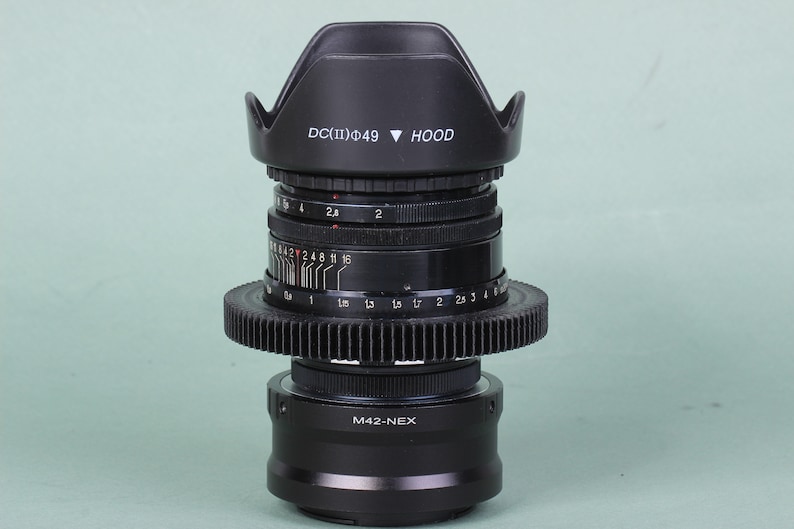 Puede incluir: Un objetivo de c&aacute;mara negro con un parasol con la inscripci&oacute;n "DC(II)&Phi;49 HOOD". El objetivo tiene varios anillos con marcas de medici&oacute;n y un anillo en forma de engranaje. La base del objetivo est&aacute; etiquetada como "M42-NEX."