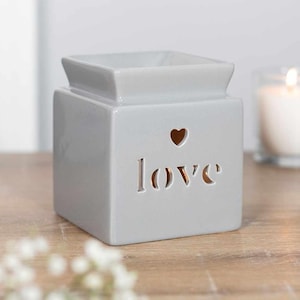 May include: A grey ceramic wax melt warmer with the word "love" cut out, and a heart-shaped cutout above the word. The warmer has a square base and a small dish on top. A lit candle is visible in the background.