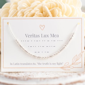 May include: A white card with a silver beaded necklace on it. The card has a heart symbol and the text "Veritas Lux Mea" in black. The text "In Latin translates to, "the truth is my light" is also on the card.