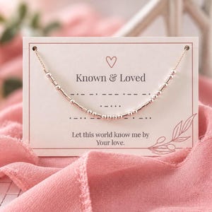May include: A rose gold-toned beaded necklace displayed on a rectangular white card. The card features a heart icon, the text Known & Loved, Morse code patterns, and the phrase Let this world know me by Your love, with a line drawing of a leaf.