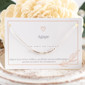 May include: A silver necklace with a small heart charm and the word "Agape" on a white card. The card also includes the definition of agape: "Highest form of love. Selfless, sacrificial, and unconditional love: persists no matter the circumstance."