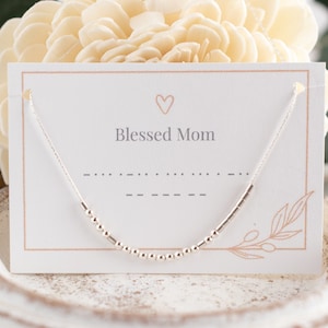 May include: A silver necklace with a beaded design and a card that reads "Blessed Mom." The card has a heart symbol and a floral design. The necklace is displayed on a white, decorative plate.