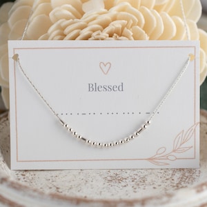May include: A silver necklace with small beads on a delicate chain. The necklace is displayed on a white card with the word "Blessed" and a heart symbol.